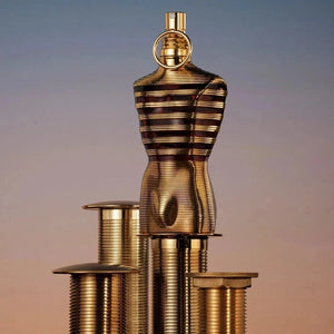 Jean Paul Gaultier Le Male Elixir Eau de Parfum bottle in a gold-striped sculpted male torso design, displayed on matching tiered industrial gold threaded pedestals against a gradient blue and orange twilight sky background.